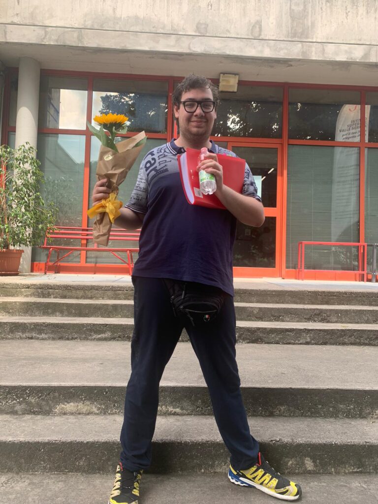 foto di Mauro all'uscita della scuola con mazzo di fiori e quaderno delle mappe dopo la prova orale, 29 giugno 2024